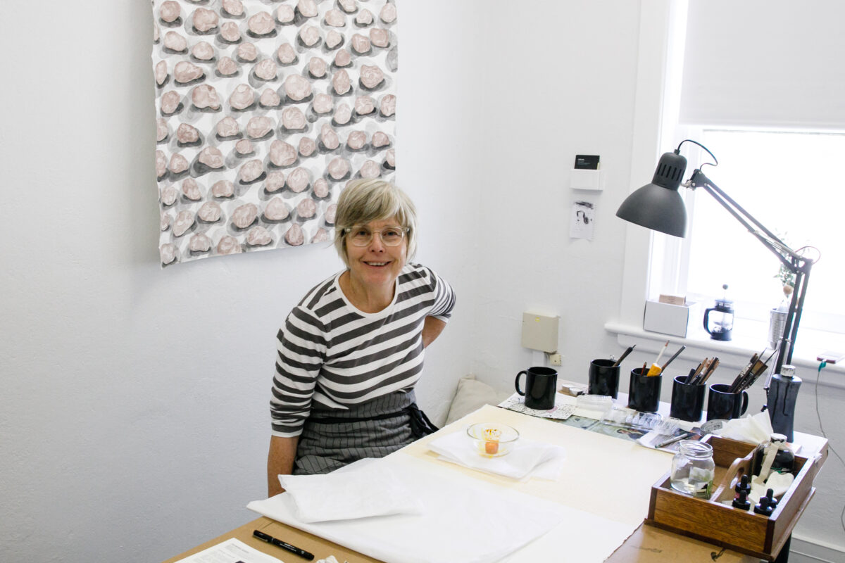 A woman wearing glasses and a striped shirt, sitting at a table with paintbrushes in mugs.