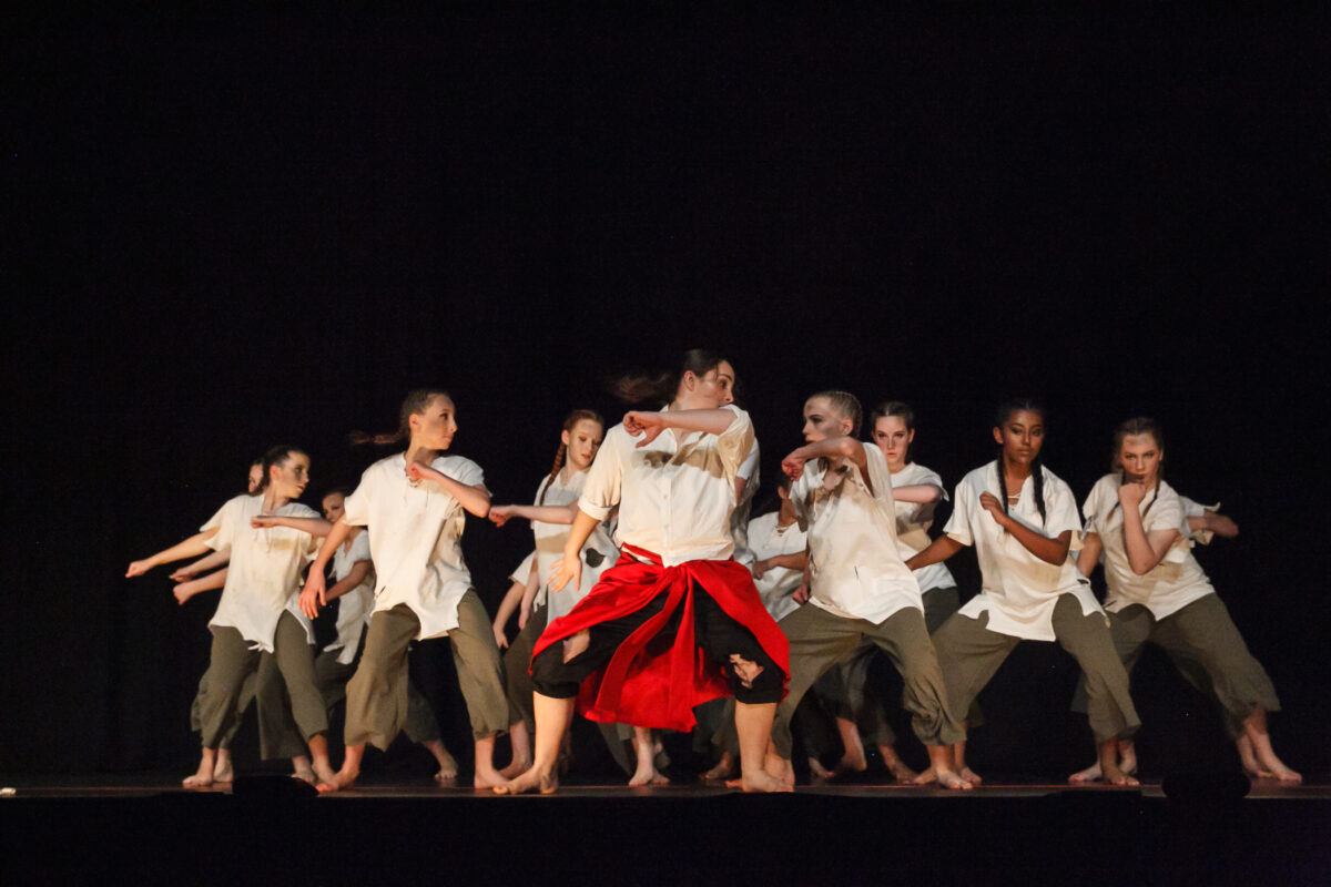 Group of women dancing on stage with a black background, wearing torn white shirts.