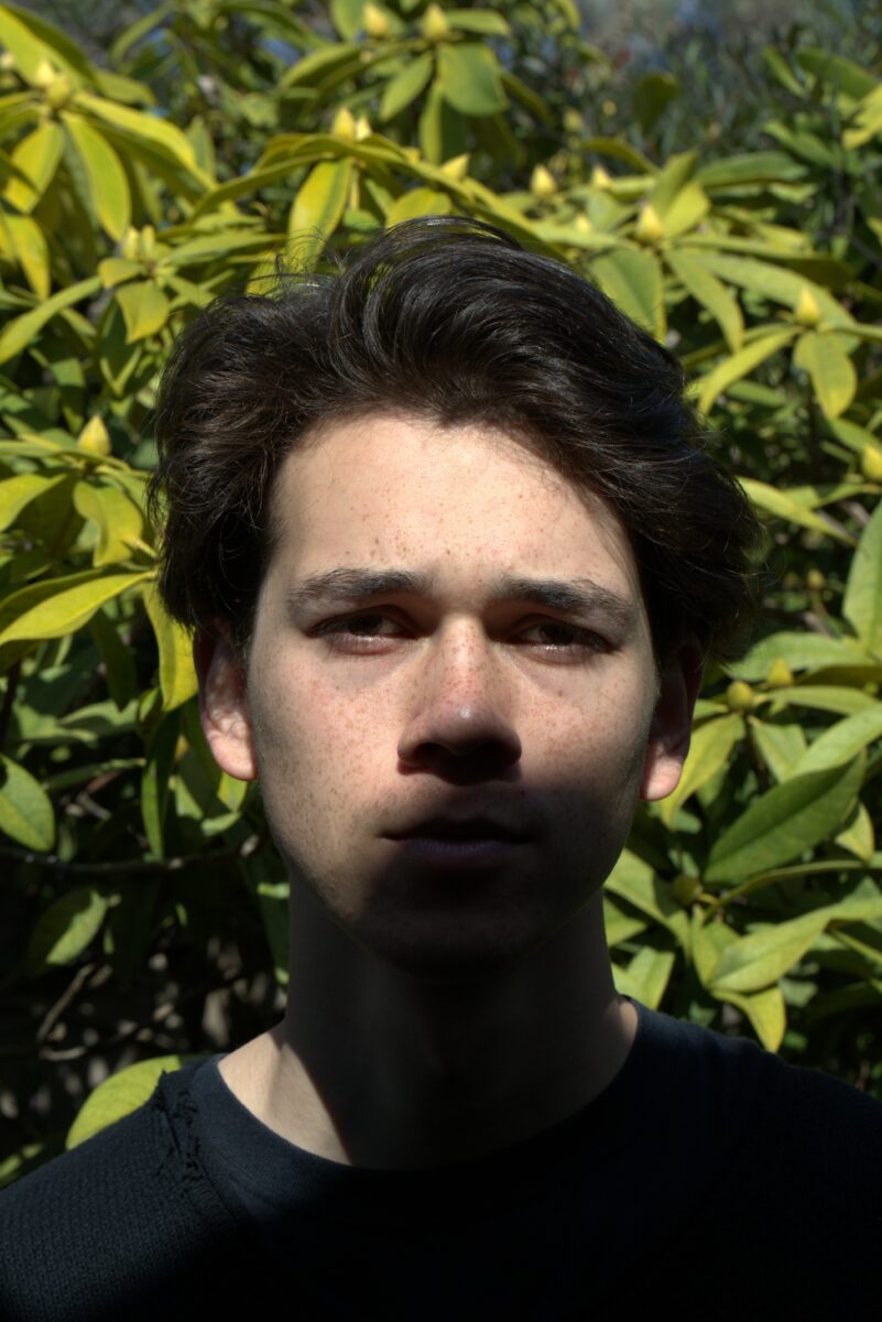 A figure stands in front of a green leafy background, the top part of their face is light by sunlight, the bottom is in the shade.