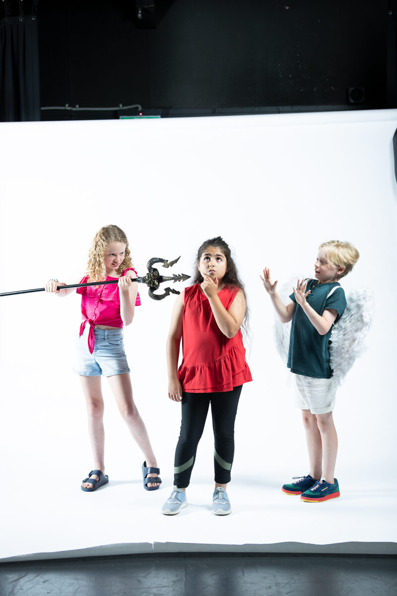 Three children stand against a white background, one dressed as a devilish character, one in the middle pondering and the third dress in angel wings.