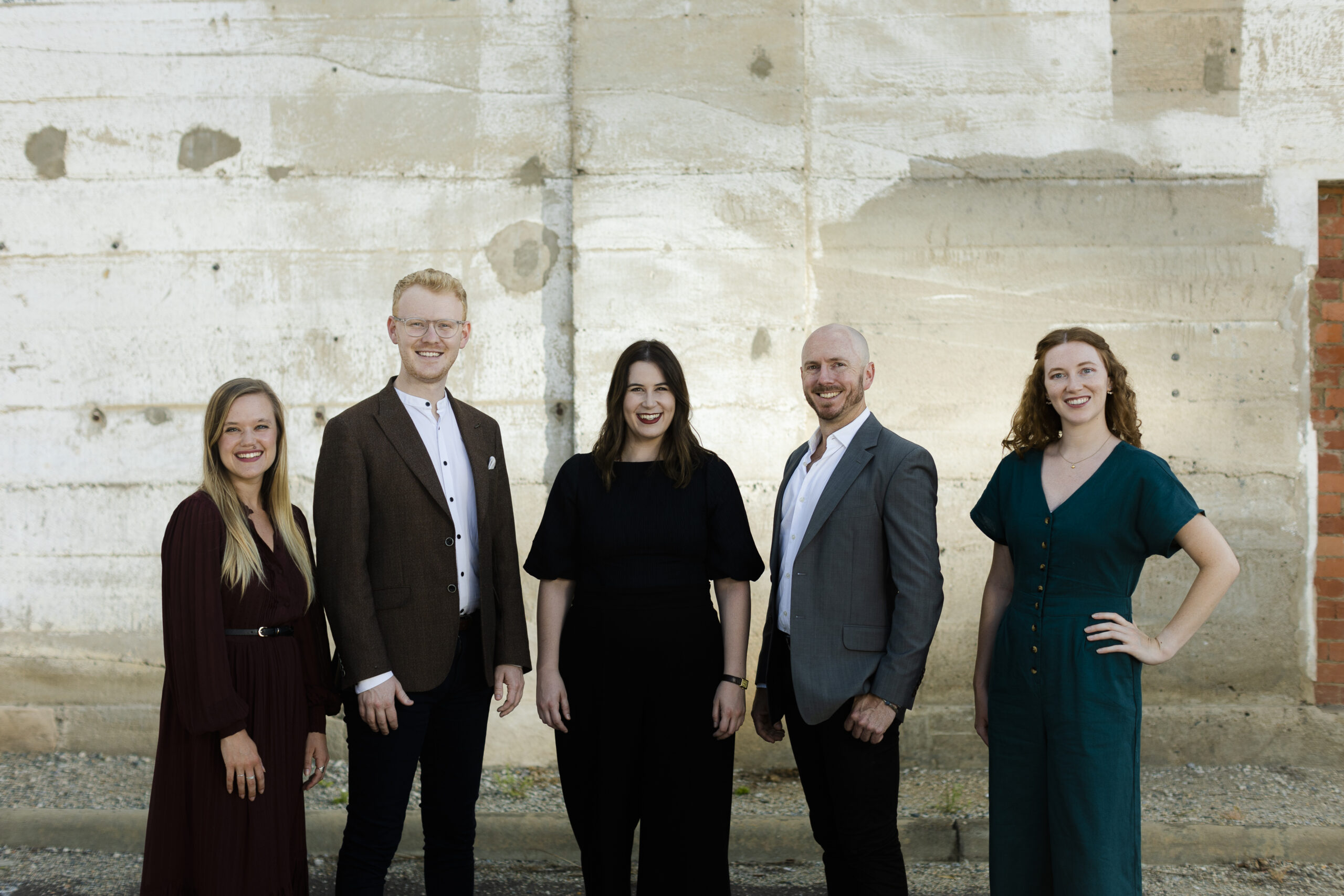 Three women adn two men stand in a line smiling at the camera. Behind them is a wall of an old building.