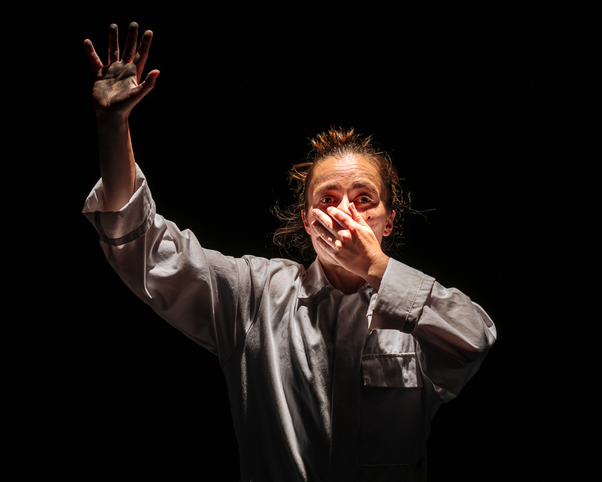 A figure in a button up shirt raises their hands in the air, one in front of their face in a dimly stage