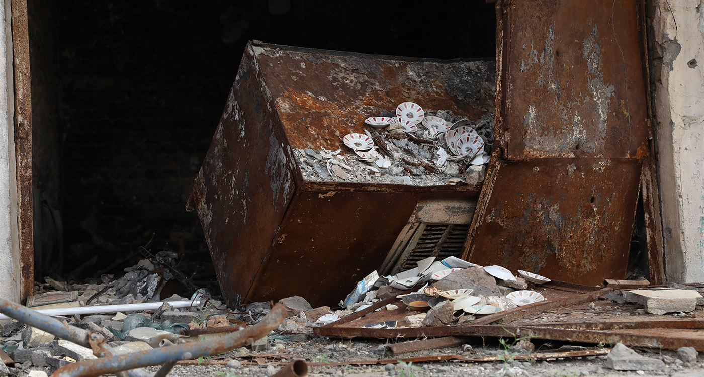 A rusted metal cart sits upturned with broken tea cup saucers. Surrounded by debris from war