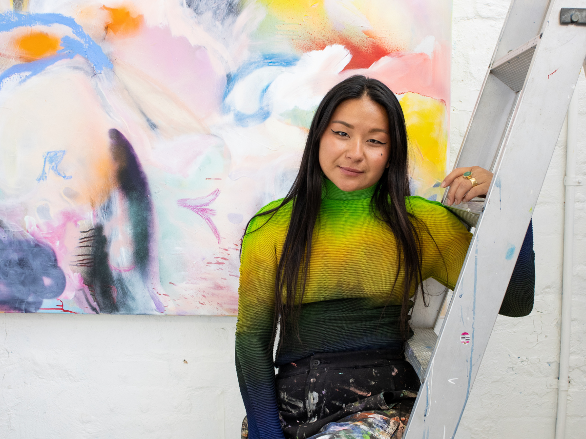 Artist Marisa Mu with black hair leans against ladder in her creative studio. One of her paintings can be seen behind her.